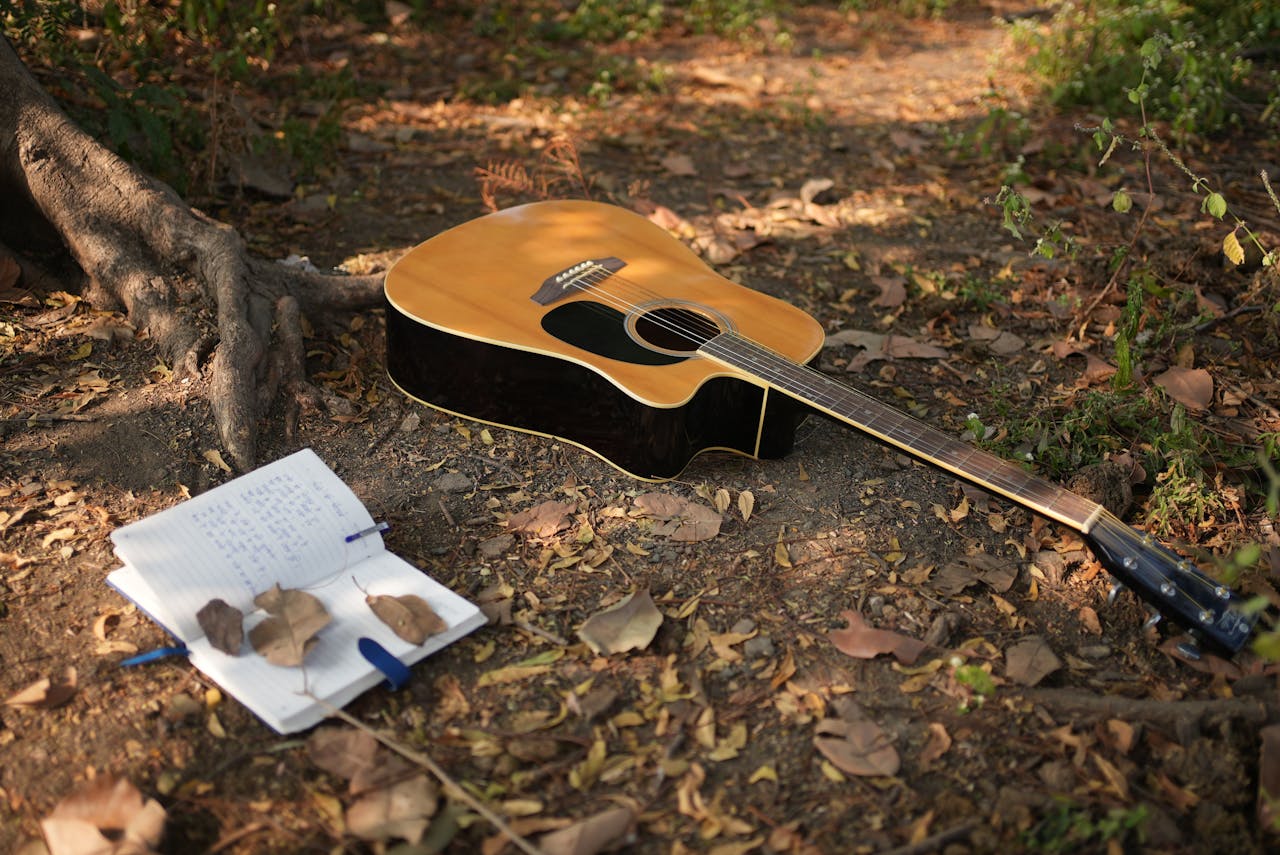 services-01 An acoustic guitar rests on forest ground beside an open notebook, capturing the essence of music creation in nature.